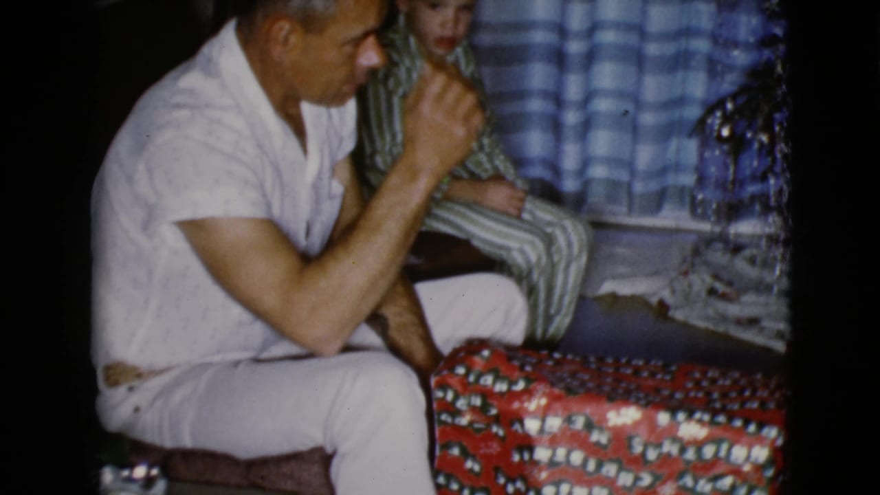 The seated man raises his hand toward his face beside the red patterned gift, suggesting surprise or emotion as the child watches in the background.