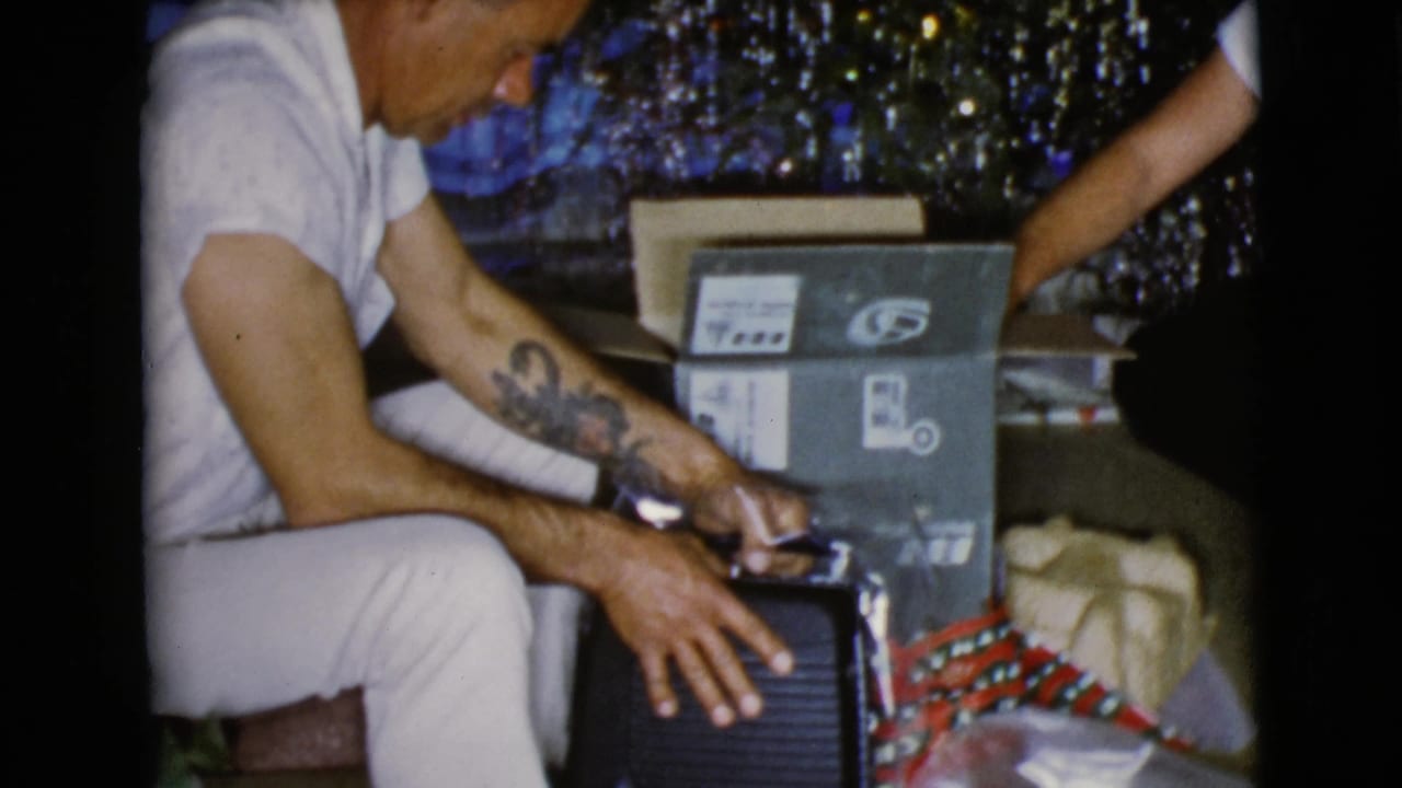 Man in a white shirt opens a present beside a tinsel Christmas tree.