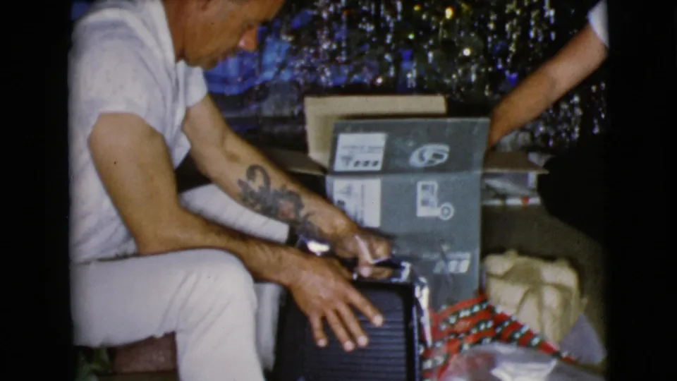 In 1968 Clarksdale, Arizona home-movie footage, a seated man opens a gift near a decorated Christmas tree and cardboard box.