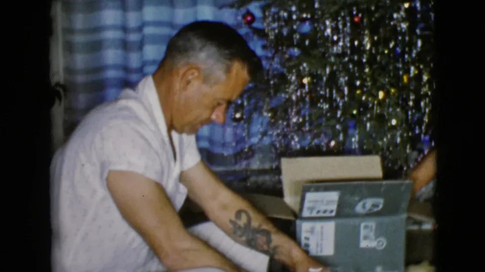 The father leans toward the present while a family member's arm reaches near a labeled box in the 1968 holiday interior.