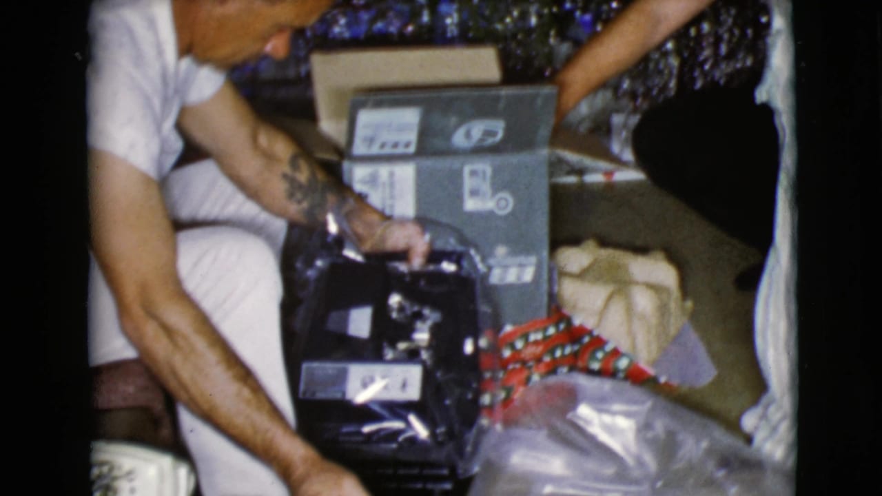 Man kneels beside wrapped gifts and opens a dark object on the floor.