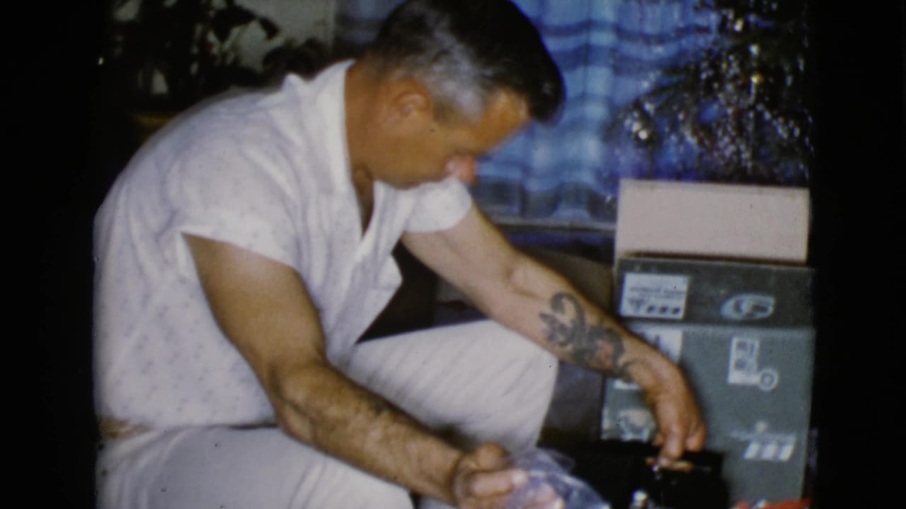 The man kneels in a private Christmas interior, reaching toward a gift while boxes and shiny decorations sit nearby.