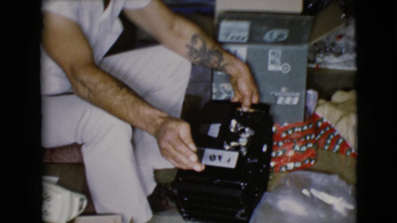 A close 1968 home-movie frame shows the man's hands holding a dark opened gift beside patterned Christmas wrapping and a box.