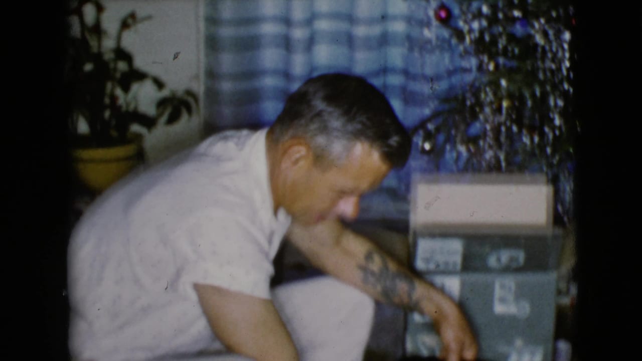 The man leans toward the floor near a tinsel Christmas tree, with blue checkered curtains and gift boxes behind him.