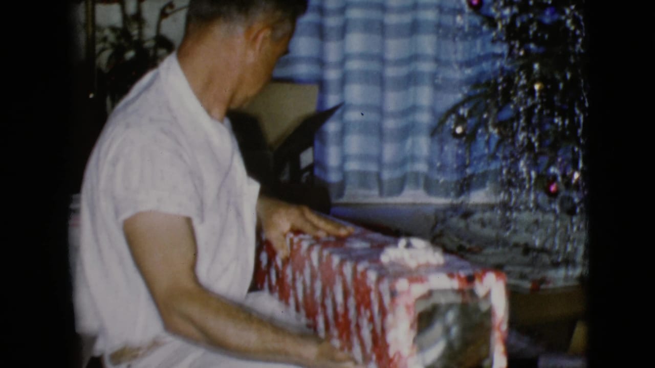 In the front room, the man turns toward a red-and-white wrapped box while the tinsel tree and checkered curtains fill the background.