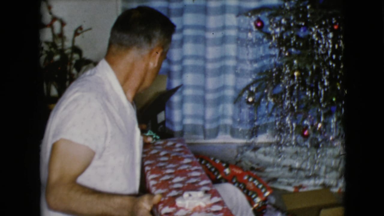 The man sits or kneels near the tree, holding a red-and-white wrapped present in the private holiday interior.