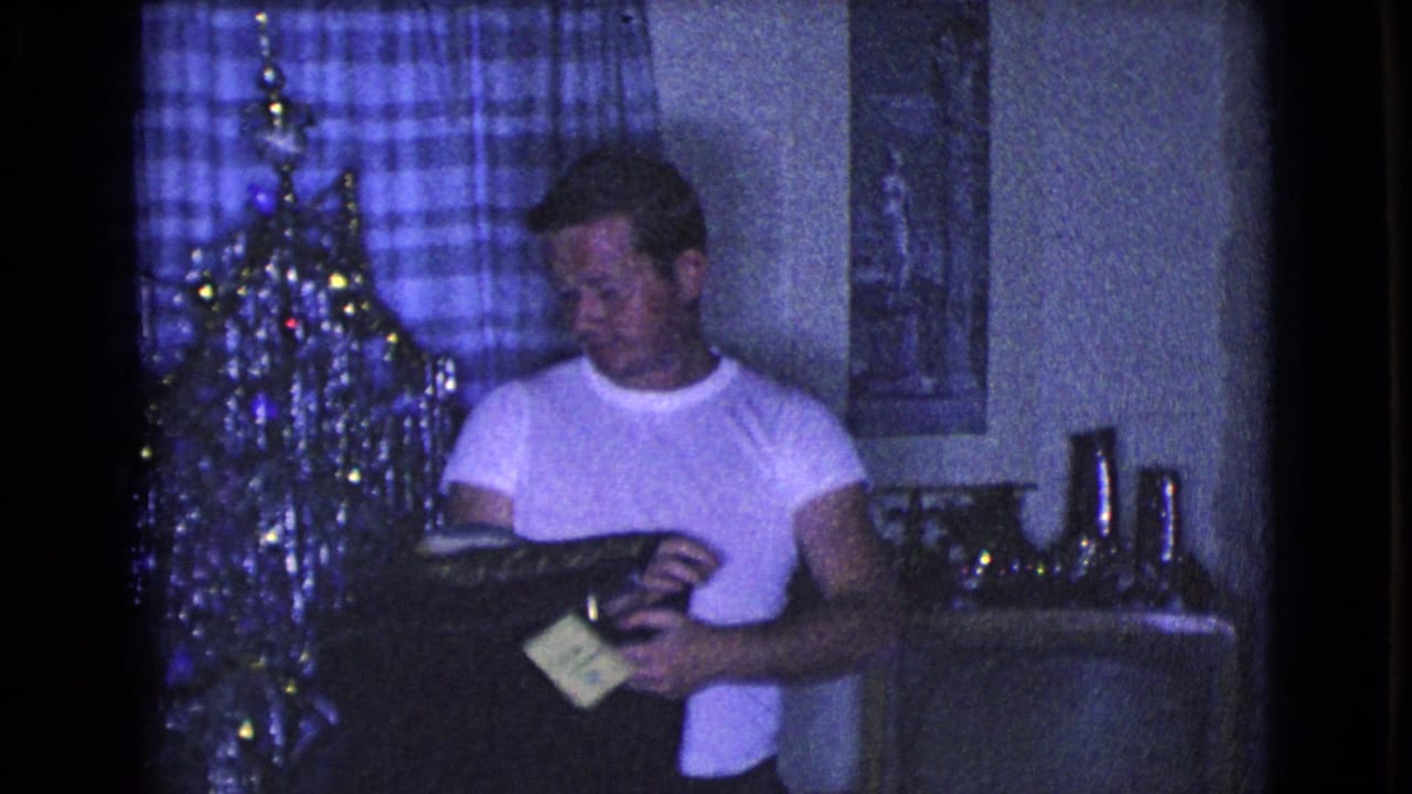 In a dim 1968 Clarksdale, Arizona living room, a man in a white shirt stands beside a Christmas tree holding a dark coat or jacket.