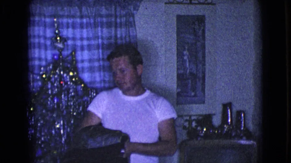 The man stands by the Christmas tree with a dark coat or jacket in hand, framed by plaid curtains and living-room decor.