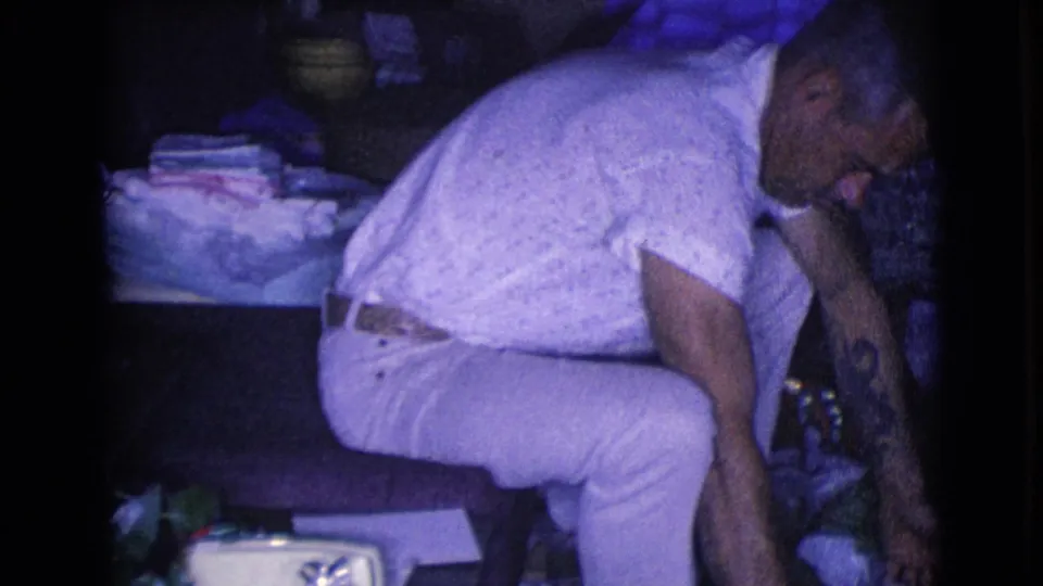 The man bends down from a chair toward packages or a box on the floor in the dim Christmas living-room setting.