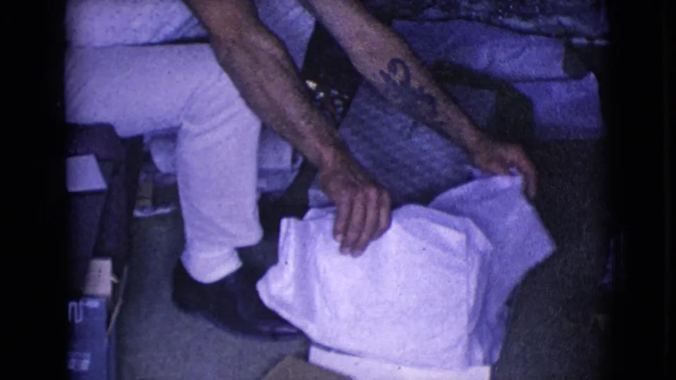 The young man remains seated in the private interior, with a textured box and loose white paper visible beside his legs.