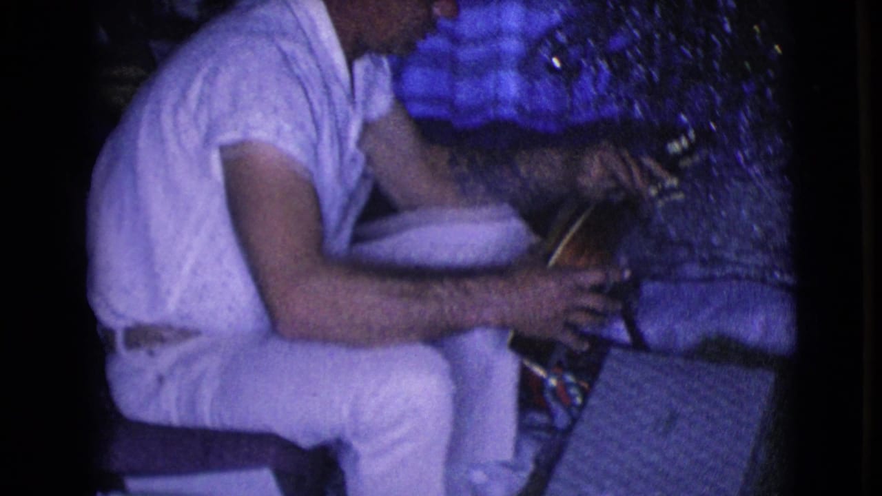 Man in white clothing seated indoors beside a Christmas tree with a small stringed instrument.