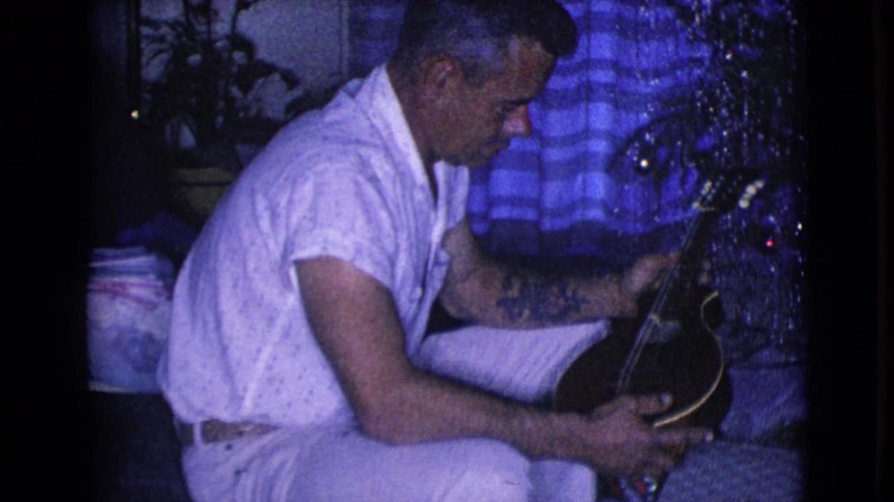 A 1968 Clarksdale, Arizona frame shows the man in profile, seated indoors and focused on a small stringed instrument near holiday decorations.