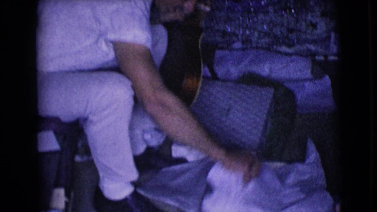 In a dim 1968 home-movie interior, a man in a light short-sleeve shirt and trousers sits near an acoustic guitar.
