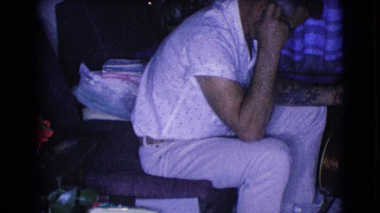Man sits sideways in a dim interior with an acoustic guitar and stacked household items nearby.