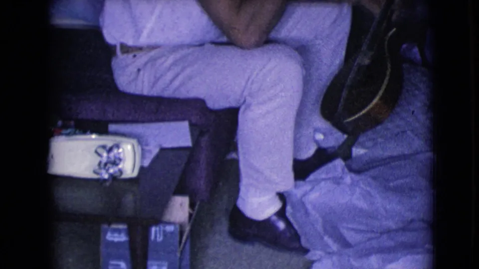 The 8mm frame shows a seated man in light clothing, an acoustic guitar, a small decorative box, and floor clutter in a private room.