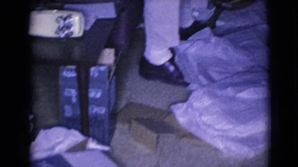 A seated man in a 1968 home-movie interior rests near an acoustic guitar, with boxes, fabric, and household objects on the floor.