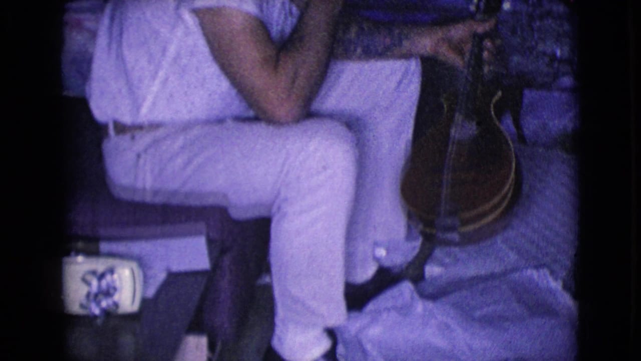 Man in light clothing seated on a couch holding a banjo in a dim living room.