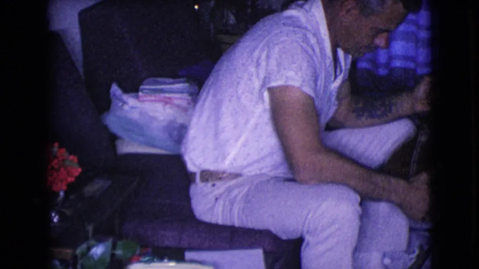1968 Clarksdale, Arizona home-movie frame with a man in profile handling a banjo near red flowers and a wrapped present.