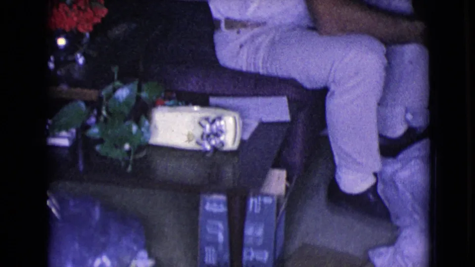 1968 8mm living-room frame from Clarksdale, Arizona with the seated man partly cropped beside red flowers and a wrapped gift.