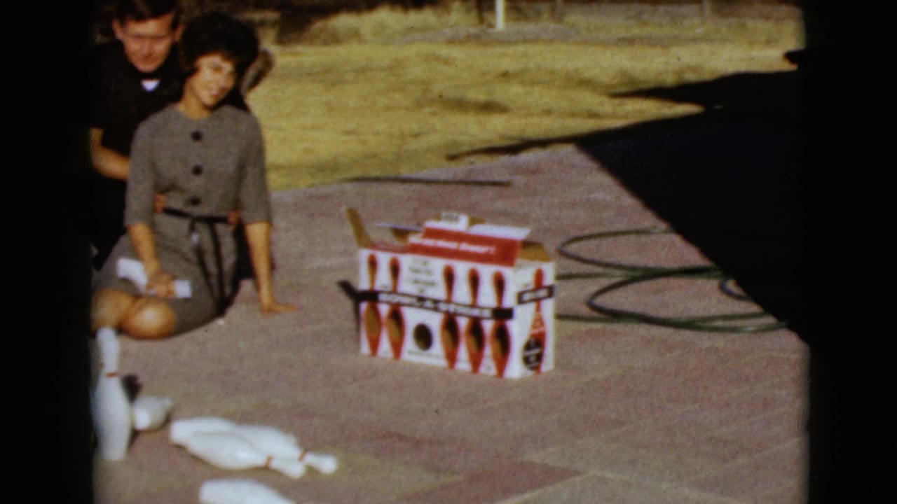 Couple on pavement beside a red-and-white mini bowling set box and scattered white pins.