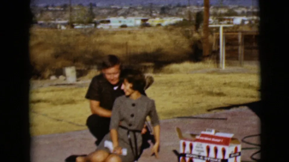 1968 8mm frame from Clarksdale, Arizona with a couple laughing near a mini bowling set, dry grass, fencing, and trailers behind them.