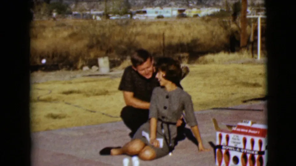 1968 Clarksdale, Arizona home-movie frame showing a smiling couple beside a mini bowling set with dry grass and trailers in the background.