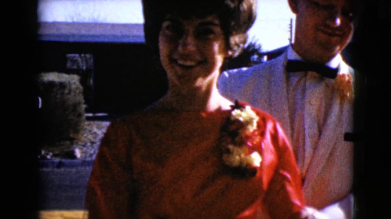 In 1968 Arizona home-movie footage, a young woman in a red dress and corsage stands with a young man in a white tuxedo jacket.