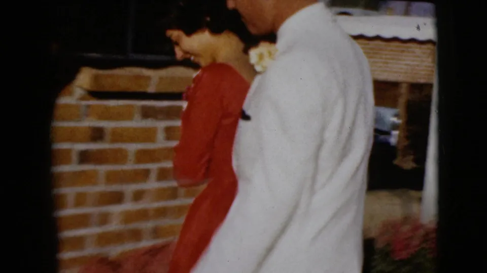 The couple walks together past a brick exterior wall, continuing the wedding-day home-movie scene in Clarksdale, Arizona.