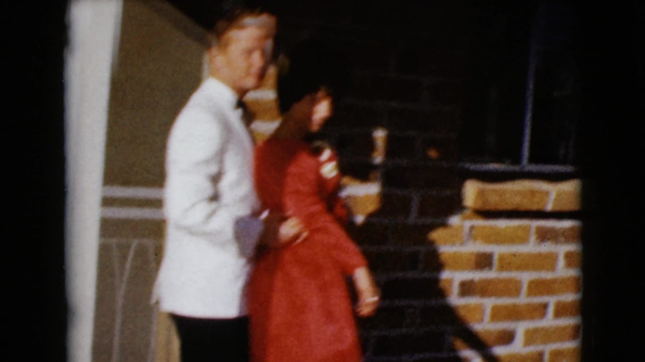 A man in a white jacket stands beside a woman in a red dress on a brick porch.