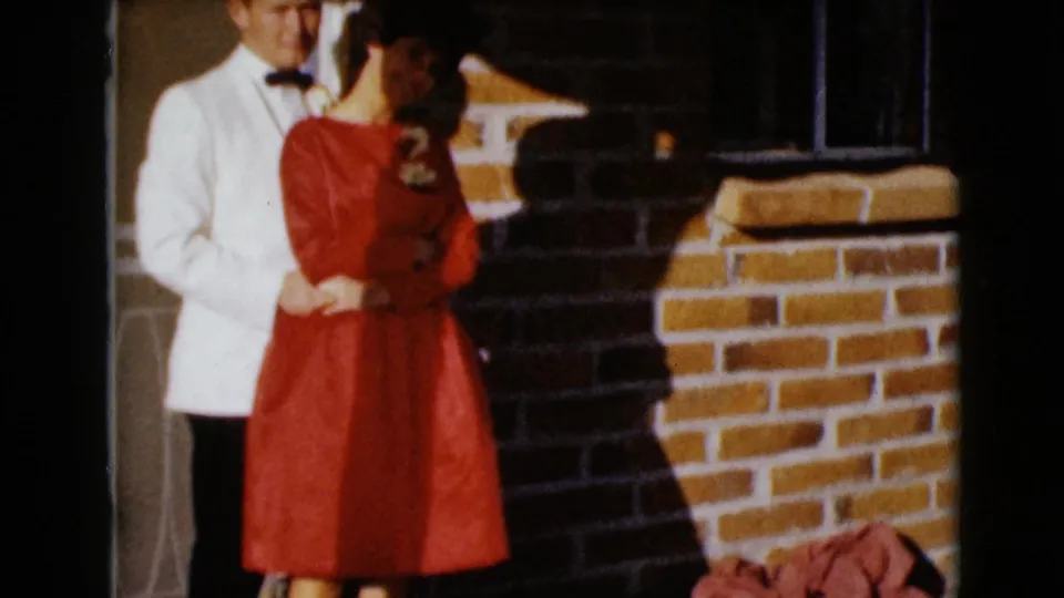 The formally dressed couple appears to adjust their pose near the doorway, with strong sunlight and shadows across the brick wall.