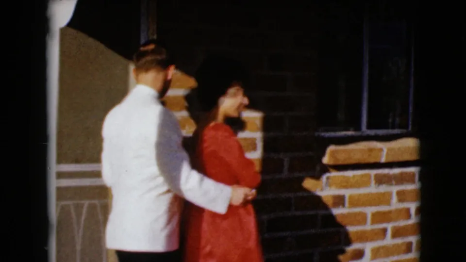A side view shows the man in a white jacket holding the woman in a red dress as they look toward the doorway area.