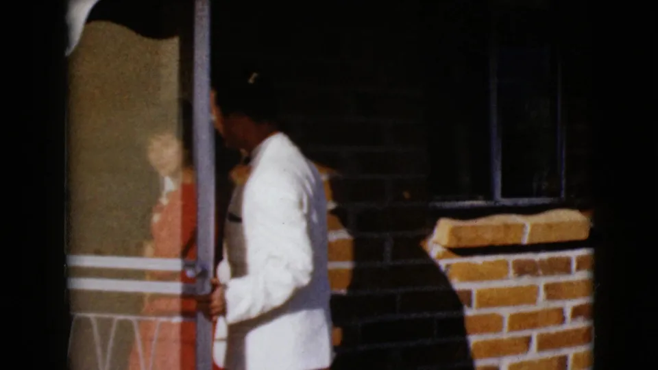 The man in formalwear holds the doorway area as the woman in red appears partly obscured behind glass or shadow near the entrance.