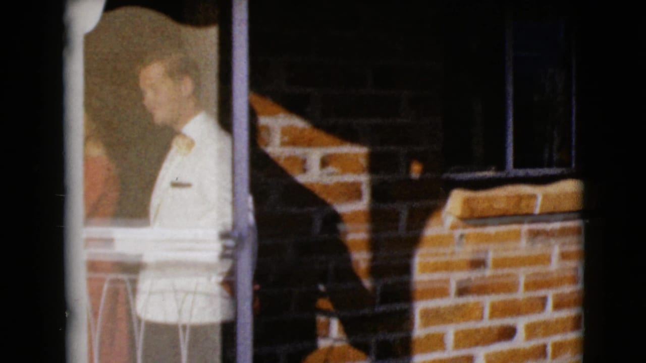 1968 8mm frame from Clarksdale, Arizona showing an adult man in a light sweater near glass and a brick residential exterior.