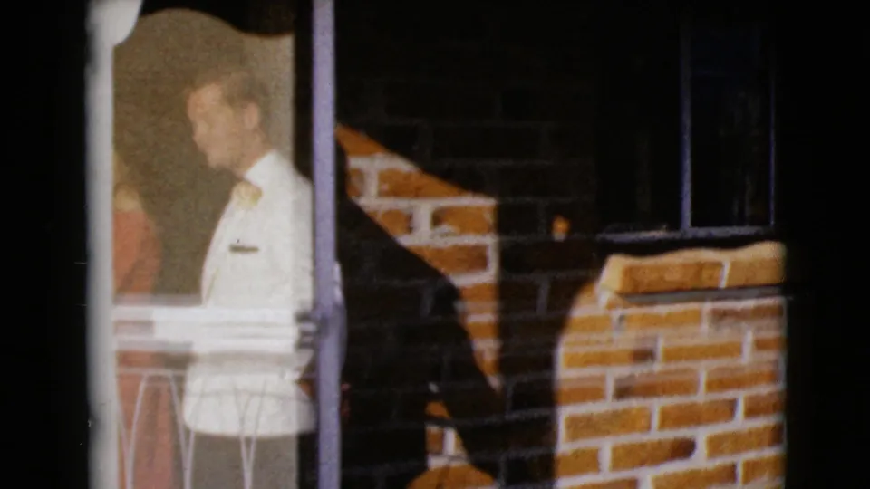 1968 8mm frame from Clarksdale, Arizona showing an adult man in a light sweater near glass and a brick residential exterior.