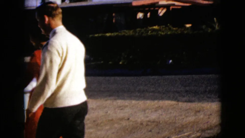 1968 Clarksdale, Arizona home-movie frame of a man and woman walking by a residential street with hedges and a parked car.