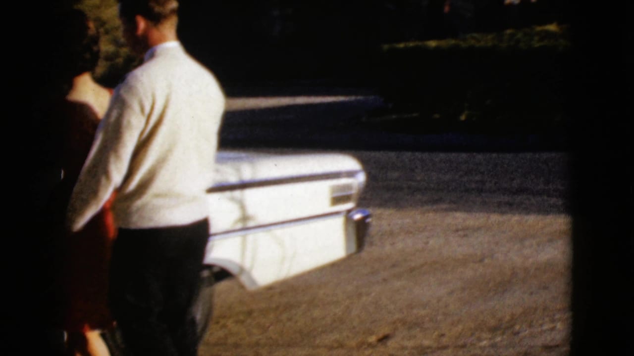 In the supplied 1968 Arizona home-movie footage, a man in a white sweater stands with a woman near the passenger side of a white sedan.