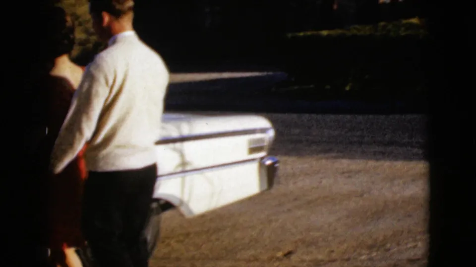 In the supplied 1968 Arizona home-movie footage, a man in a white sweater stands with a woman near the passenger side of a white sedan.