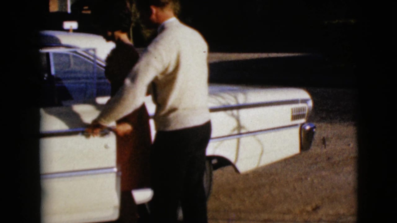 The man appears to hold or steady the passenger door area while the woman stands beside the white 1960s sedan in daylight.
