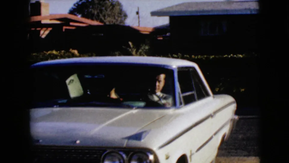A white 1960s sedan is seen from the front in a residential setting, with an adult visible through the windshield.