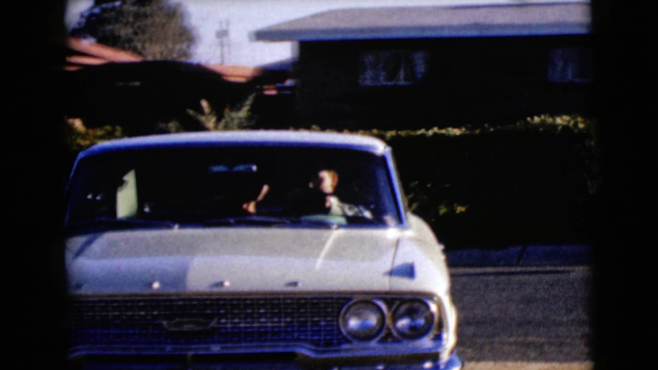 White vintage sedan seen head-on in a driveway with two occupants visible through the windshield.