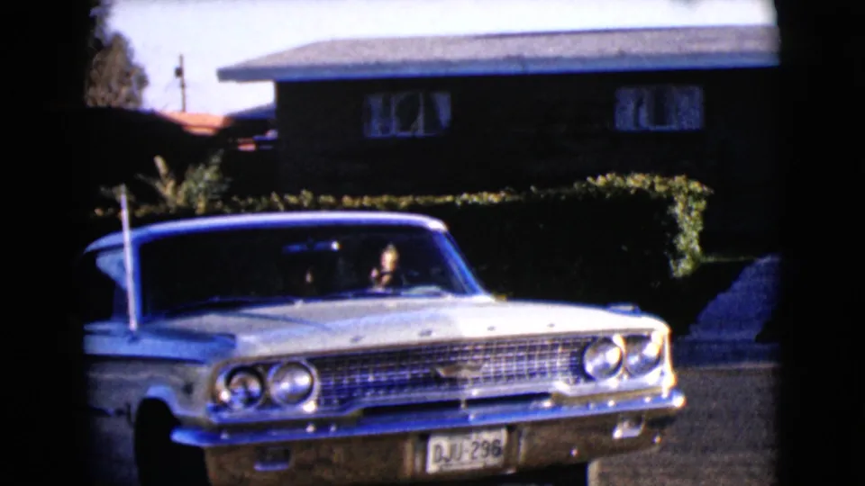 1968 Clarksdale, Arizona frame of a white classic sedan backing or turning from a driveway, with the driver visible and the front plate readable.