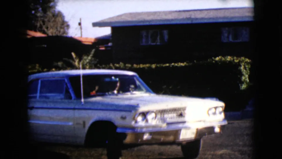 1968 Clarksdale, Arizona home-movie frame showing the white sedan from a front-side angle beside a hedge and dark residential house.