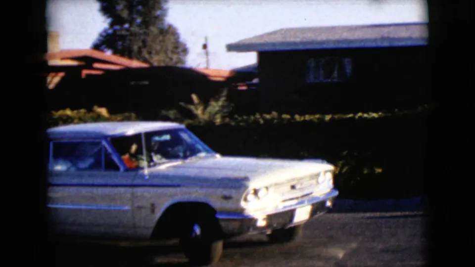 1968 Clarksdale, Arizona frame of the white sedan continuing away from the camera beside a hedge and suburban house, with occupants faintly visible.