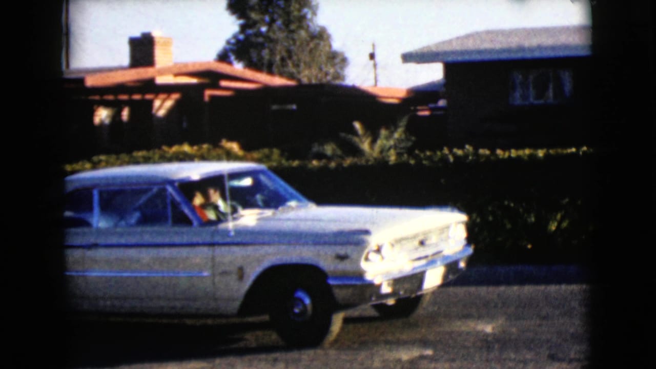 The white sedan continues past hedges and low houses, with at least one person visible inside the car in the 1968 Arizona home-movie frame.