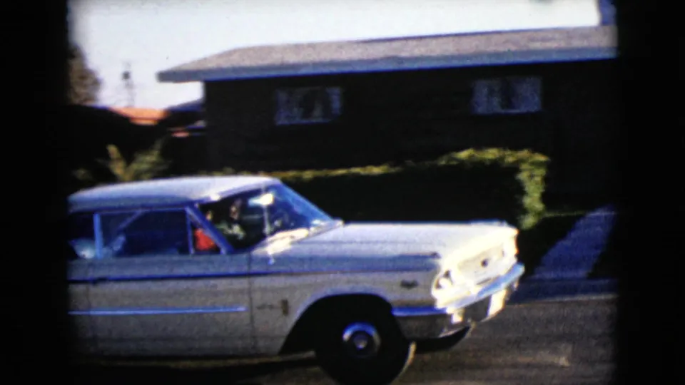 A white mid-century sedan passes a hedge-lined house on a bright residential street in 1968 Clarksdale, Arizona footage.