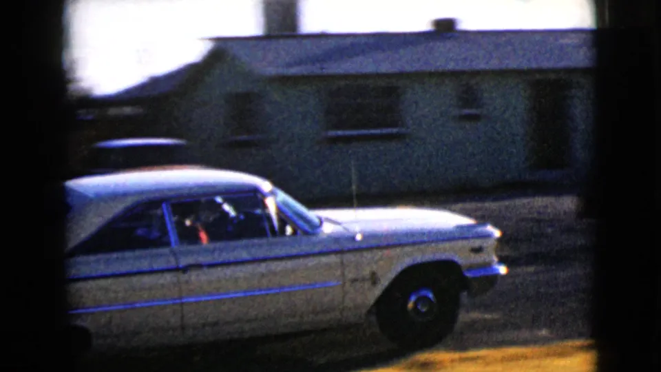 The white sedan is shown in side view near a low residential house, with dark 8mm borders and strong sunlight in the frame.