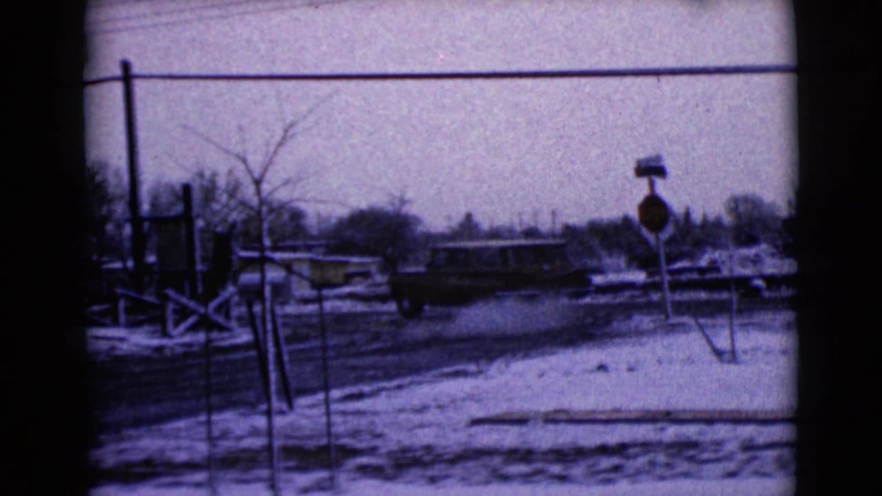 Snowy street with a light-colored car passing a stop sign and roadside poles.