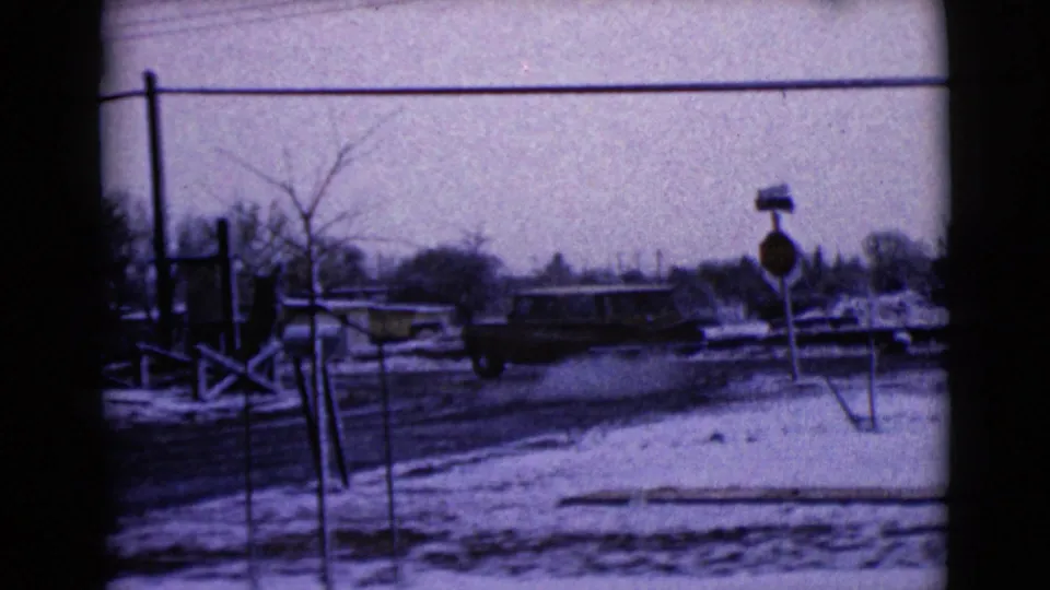 1968 Clarksdale, Arizona frame with a light-colored car crossing a snowy residential street past signs, poles, and bare trees.