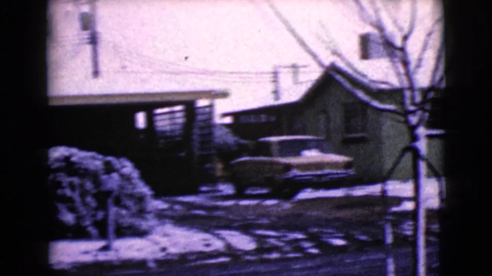1968 home-movie frame showing a classic car parked in a snowy driveway among low residential buildings in Clarksdale, Arizona.
