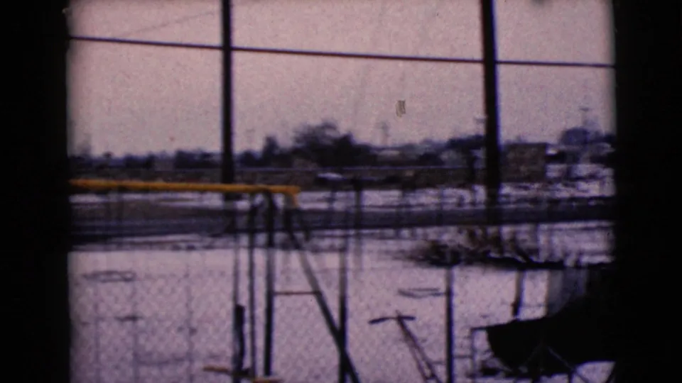 1968 8mm frame in Clarksdale, Arizona, looking across a fenced snowy playground toward poles, wires, and low distant structures.